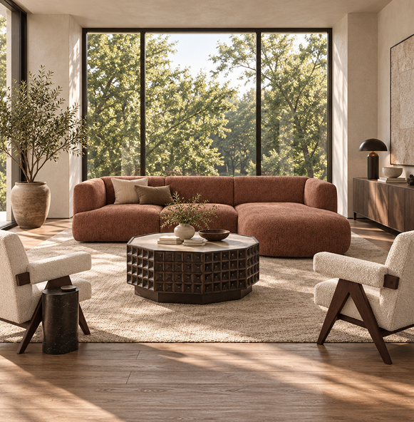 A modern living room featuring a large pink modular sectional sofa, a dark geometric coffee table, two cream armchairs, and a large beige rug, set against a background of tall windows showing a lush green outdoor view.